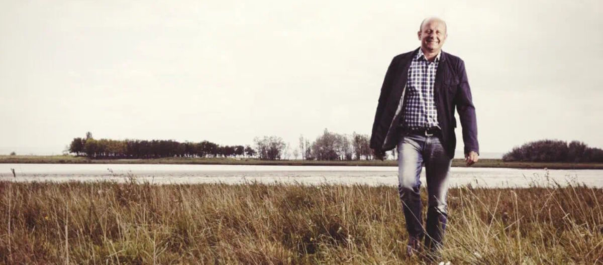 Mann in Freizeitkleidung geht durch eine offene Wiesenlandschaft mit Wasser im Hintergrund und leicht bewölktem Himmel