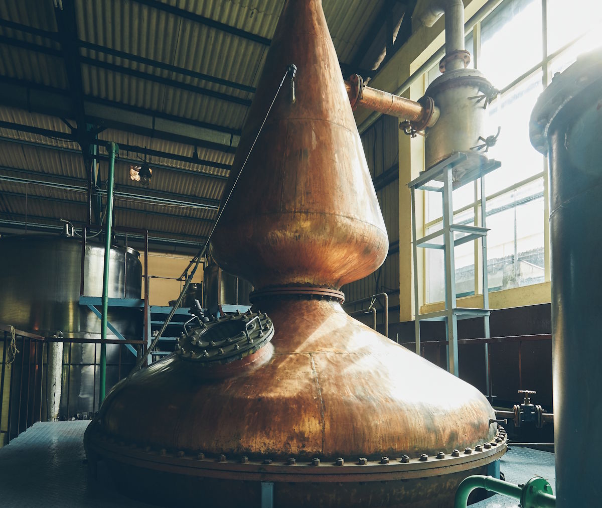 Große kupferne Brennblase in einer industriellen Destilleriehalle mit hohen Fenstern und technischer Ausstattung – klassisches Destillationsgerät für die Herstellung von Whisky