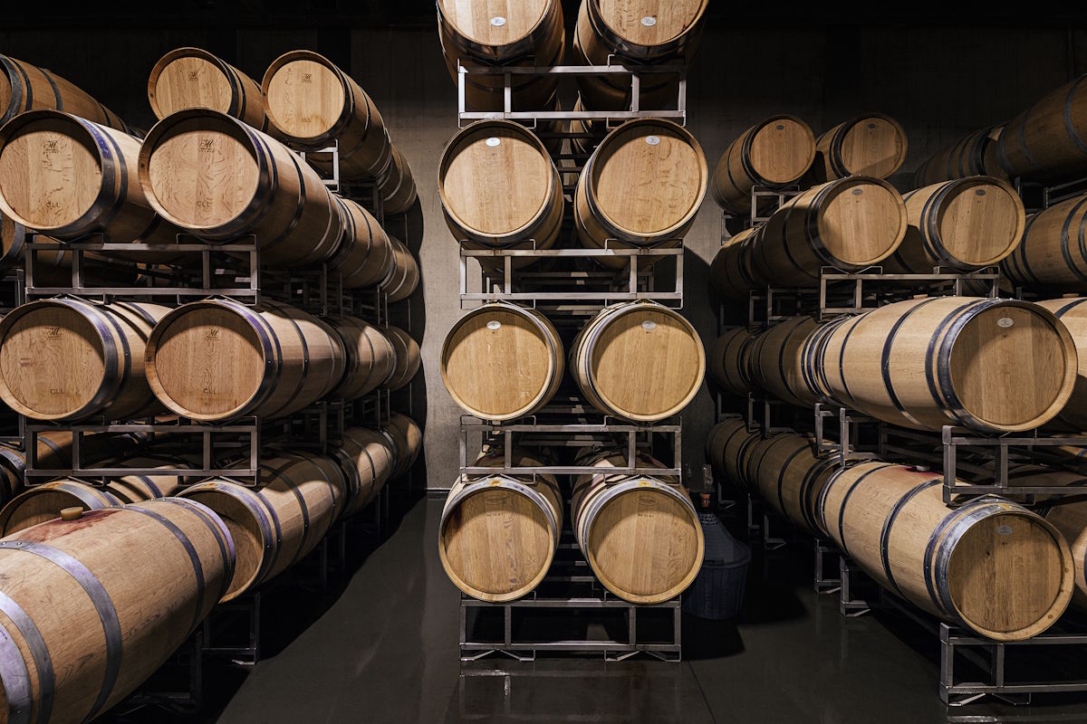 Gestapelte Eichenfässer zur Weinreifung in einem modernen Keller mit Metallgestellen und dunklem Boden.