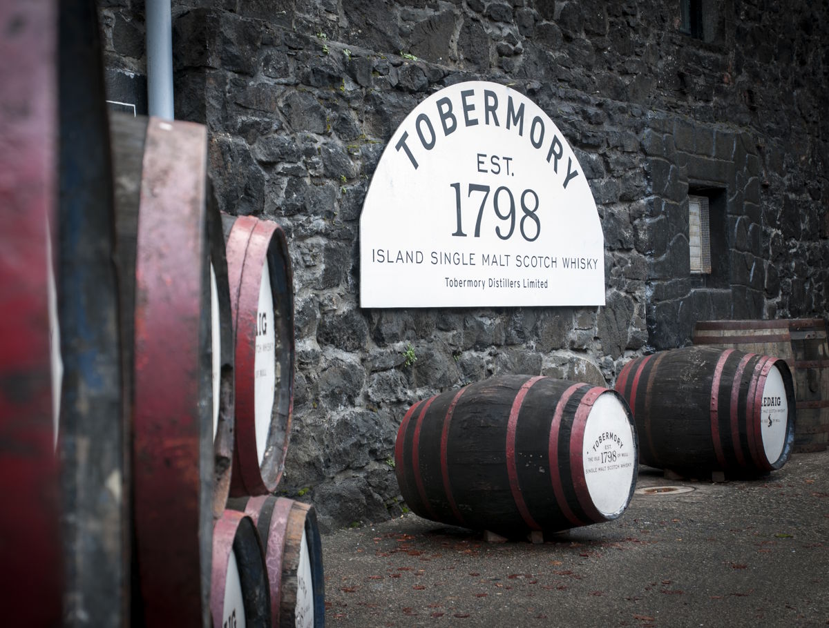 Historische Fassade der Tobermory Distillery mit einem Schild „Tobermory Est. 1798 – Island Single Malt Scotch Whisky“ an einer Steinmauer und mehreren rot-schwarz lackierten Whiskyfässern davor