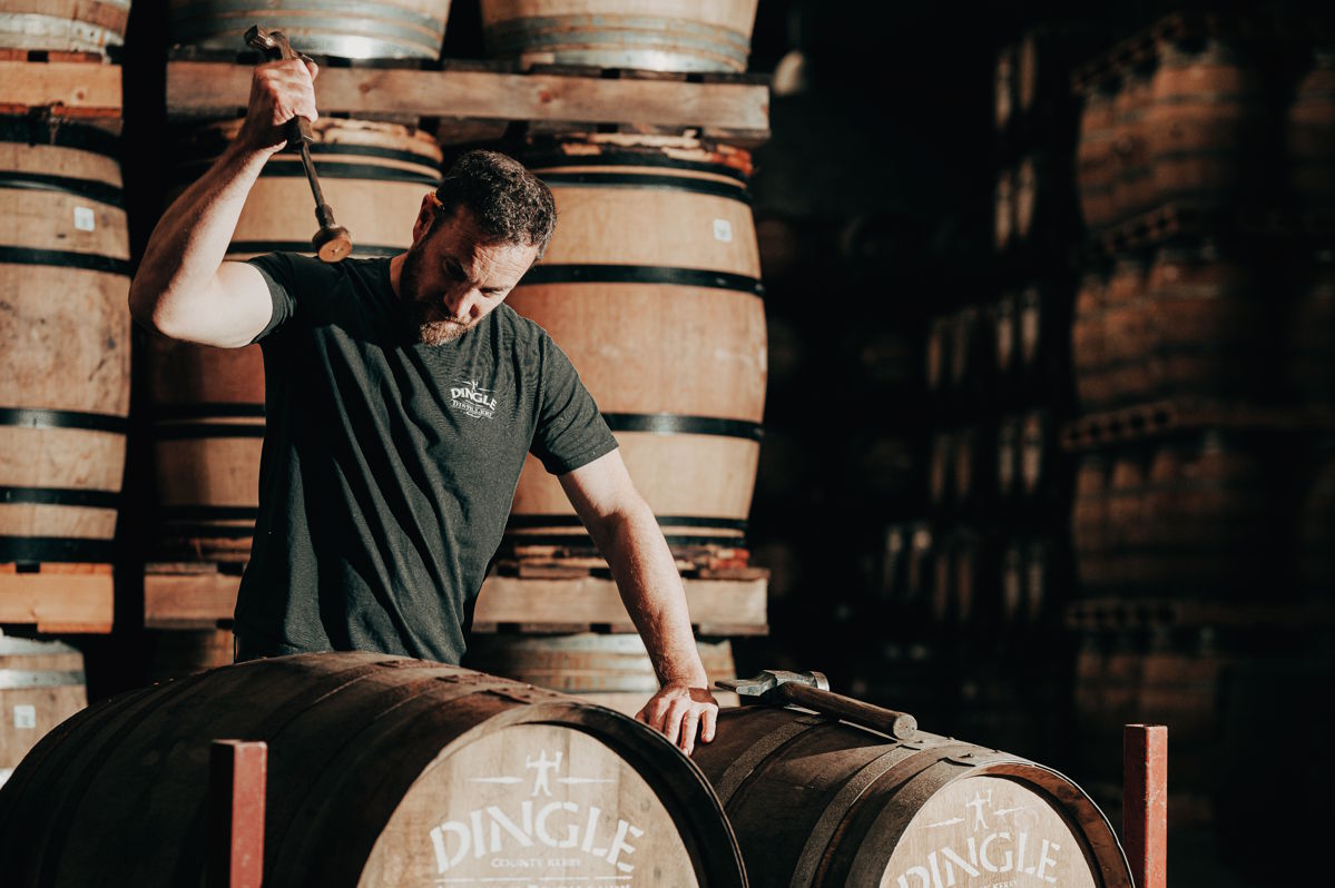 Mitarbeiter einer Whisky-Destillerie schlägt mit einem Werkzeug auf ein Eichenfass, um es zu verschließen – im Hintergrund zahlreiche gelagerte Holzfässer in einem traditionellen Lagerhaus mit dunkler Atmosphäre