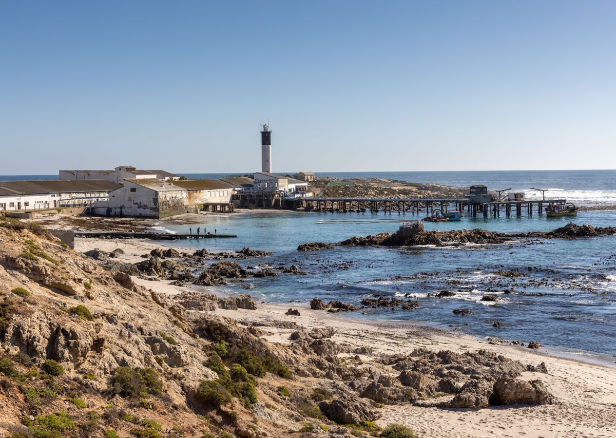 Küstenlandschaft mit Felsen, Sandstrand und einem historischen Pier, im Hintergrund ein Leuchtturm und alte Gebäude direkt am Meer.
