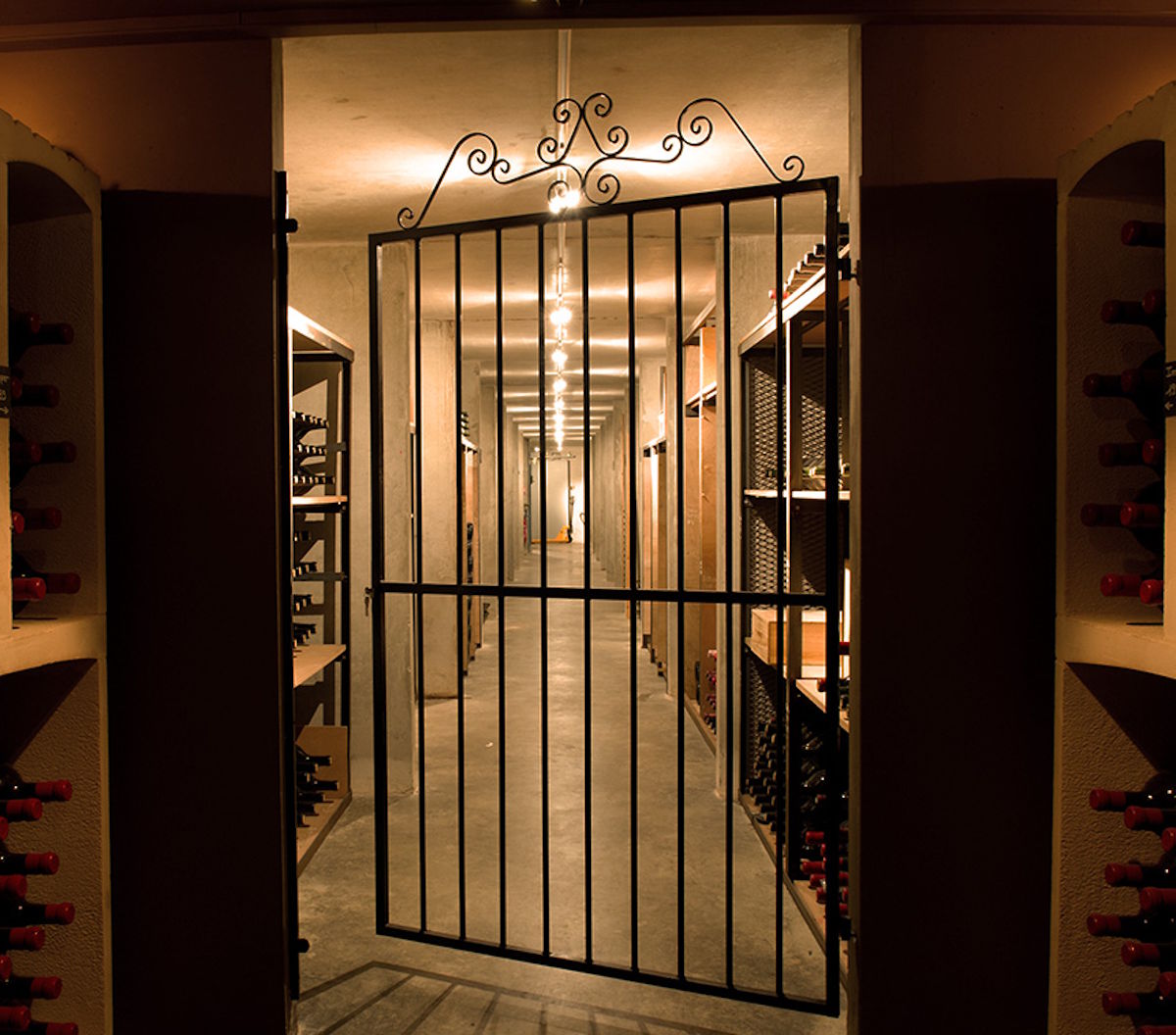 Blick durch ein schmiedeeisernes Tor in einen stimmungsvoll beleuchteten Weinkeller mit Regalreihen voller Weinflaschen.