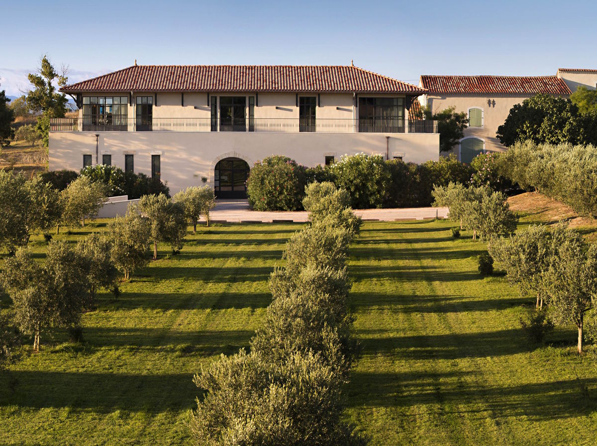 Mediterranes Weingut mit rotem Ziegeldach und gepflegtem Garten, in dem symmetrisch gepflanzte Olivenbäume in der Nachmittagssonne Schatten werfen.