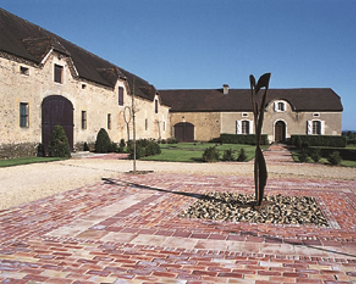 Historische Gebäude eines Weinguts mit hellem Steinmauerwerk und dunklen Holztoren, umgeben von gepflegten Wegen, Rasenflächen und einer modernen Metallskulptur im Hof.