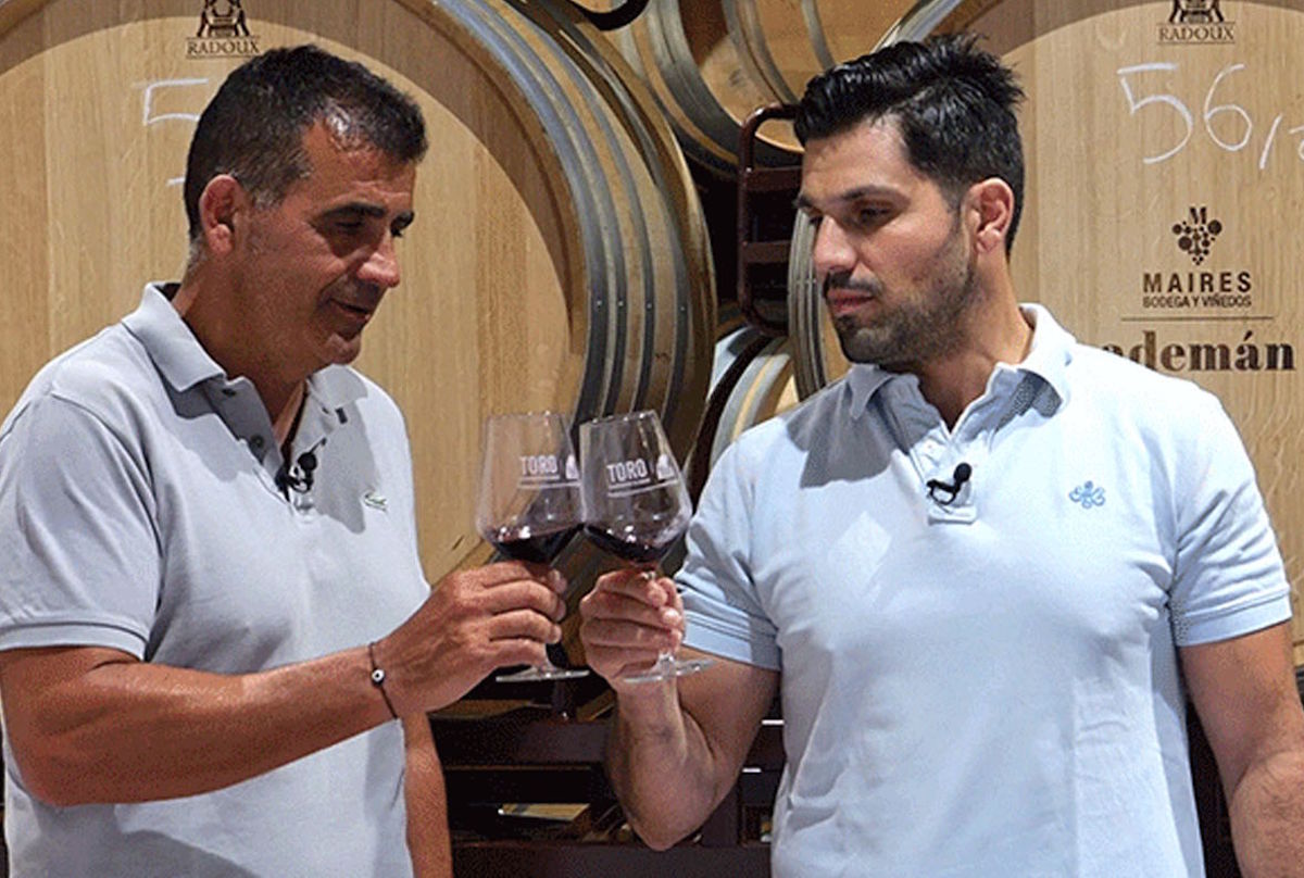 Zwei Männer stoßen in einem Weinkeller mit Rotweingläsern an, im Hintergrund große Holzfässer mit Prägung