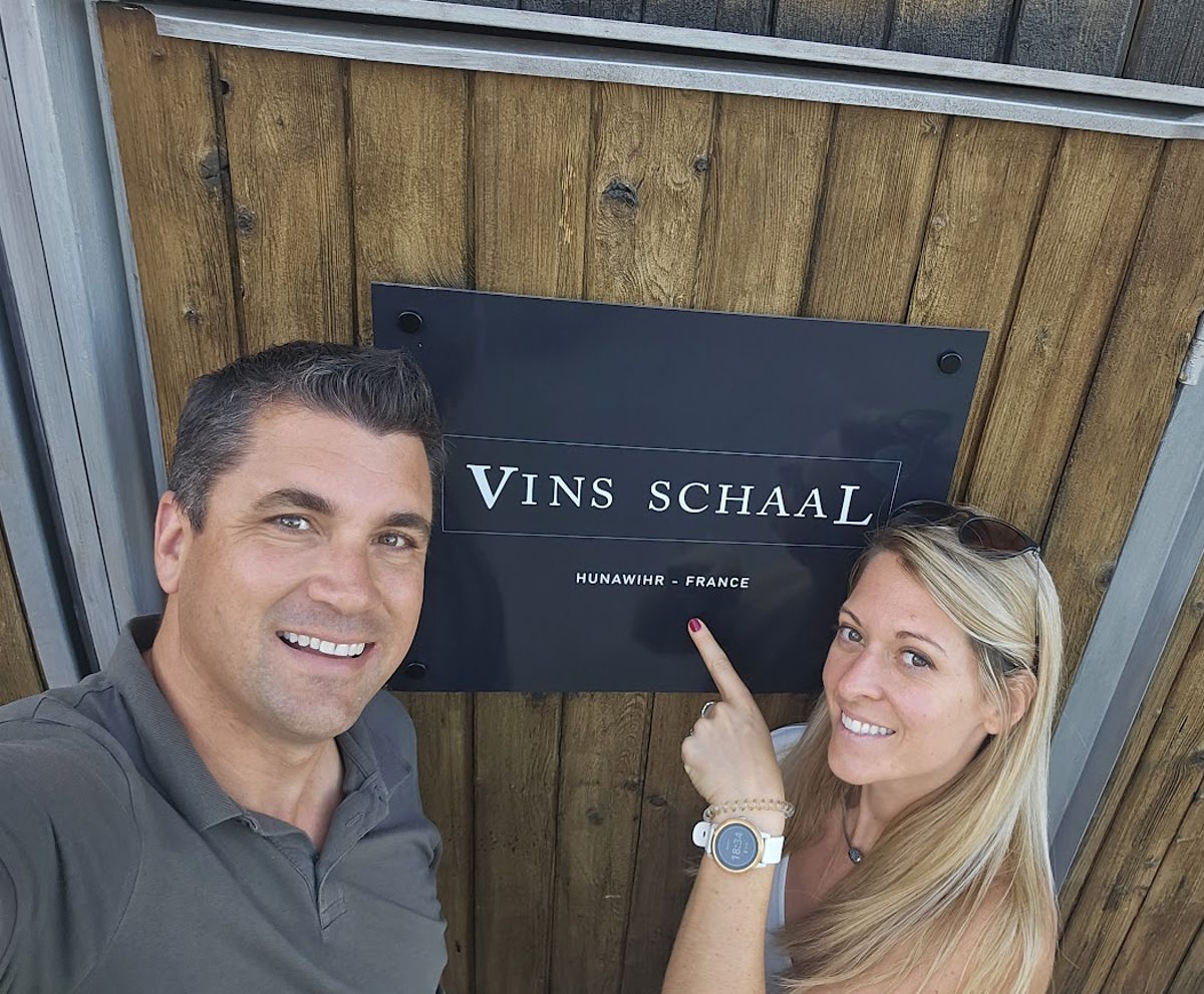 Ein Mann und eine Frau lächeln vor einem Schild mit der Aufschrift „Vins Schaal – Hunawihr, France“ an einer Holzwand.
