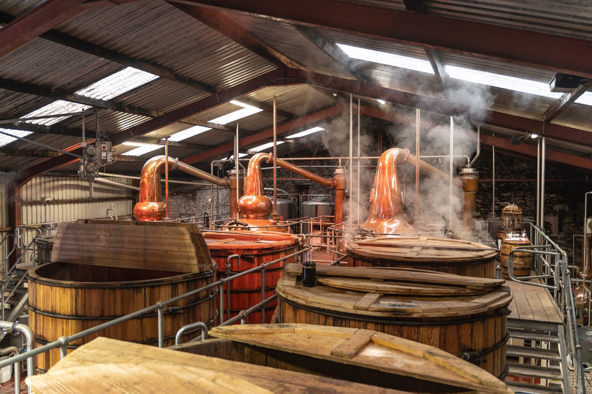 Kupferbrennblasen und hölzerne Gärbottiche in einer traditionellen Whisky-Brennerei mit Blechdach – authentischer Einblick in den Herstellungsprozess von Single Malt Whisky