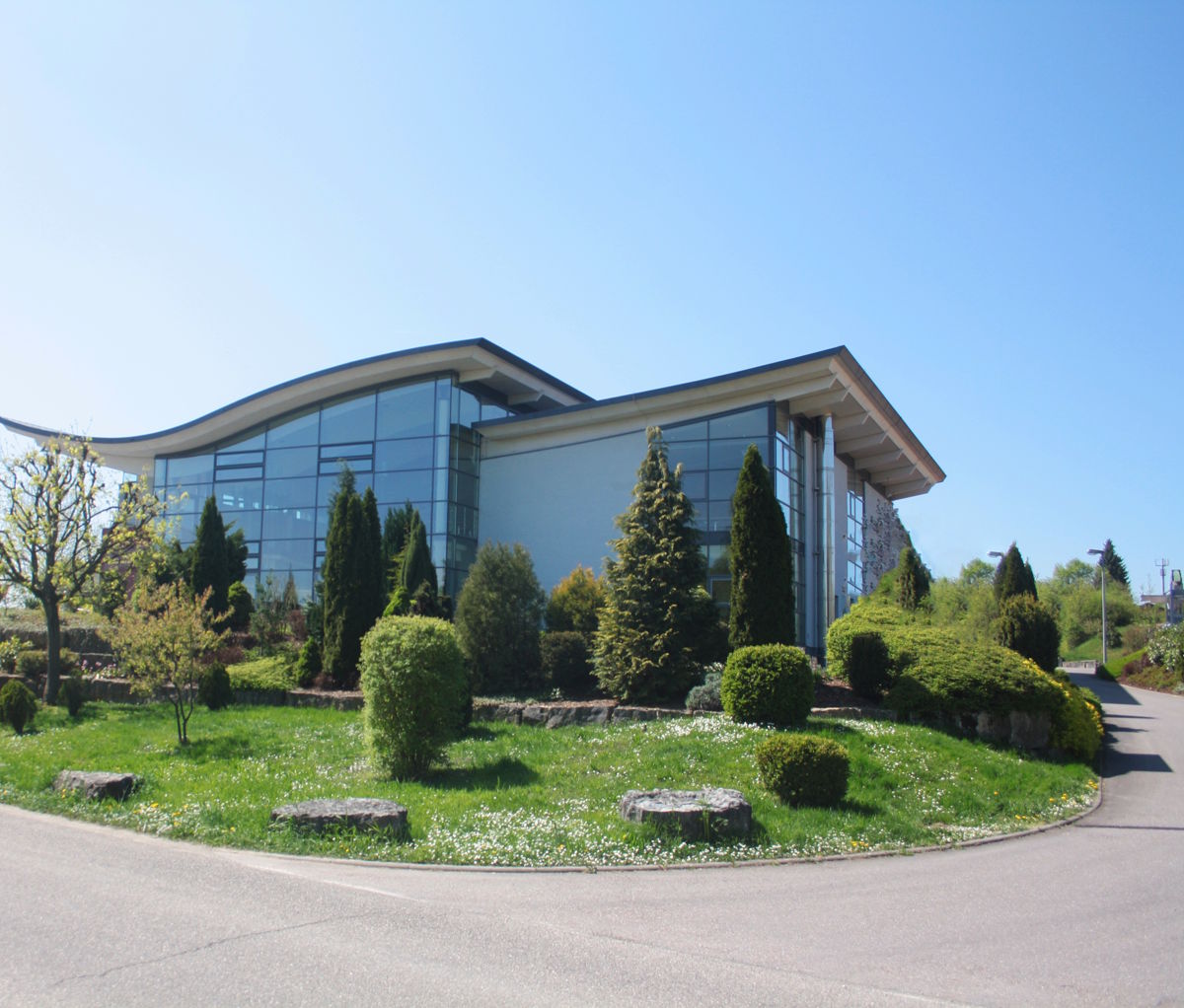 Modernes Weingutsgebäude mit großer Glasfassade, umgeben von gepflegtem Garten und Bäumen bei sonnigem Wetter