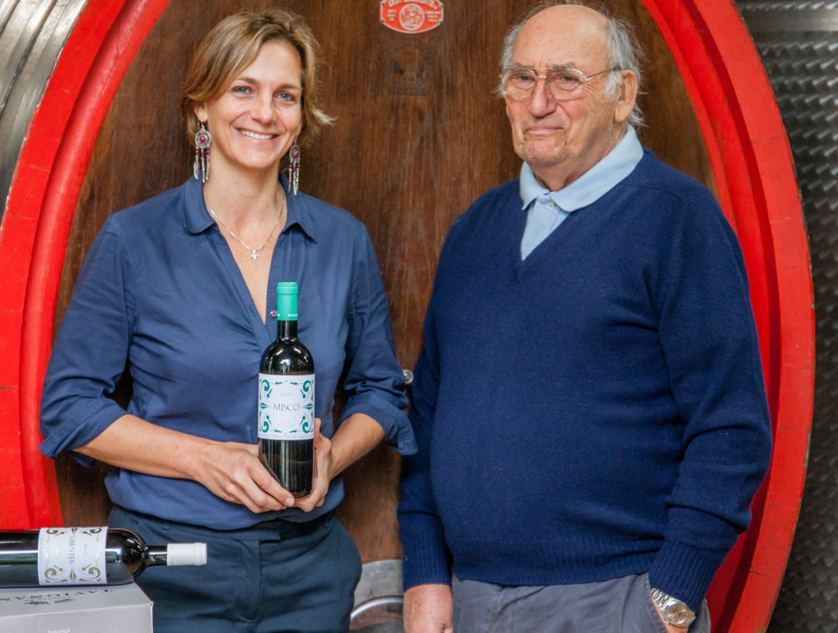 Winzerin und älterer Winzer stehen lächelnd vor einem Holzfass und präsentieren eine Flasche Wein