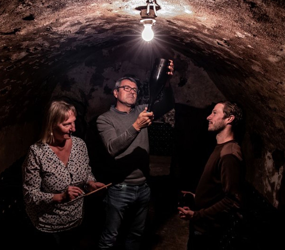 Drei Personen in einem alten, gewölbten Weinkeller, eine Flasche wird gegen das Licht gehalten und begutachtet.