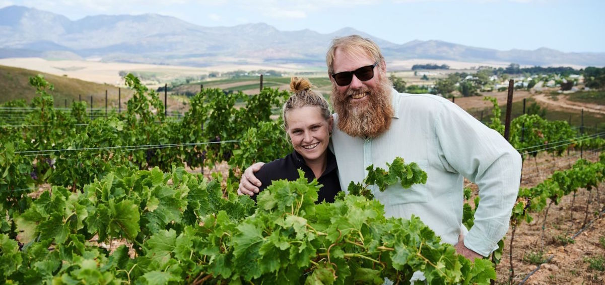 Winzerpaar steht lächelnd zwischen Reben auf einem Hügel, im Hintergrund Weinberge und Berge unter blauem Himmel.