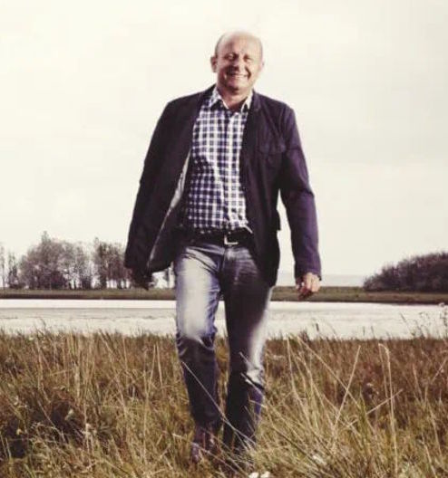 Fröhlich lächelnder Mann in kariertem Hemd und Blazer spaziert durch eine Wiese vor einem See oder Flussufer