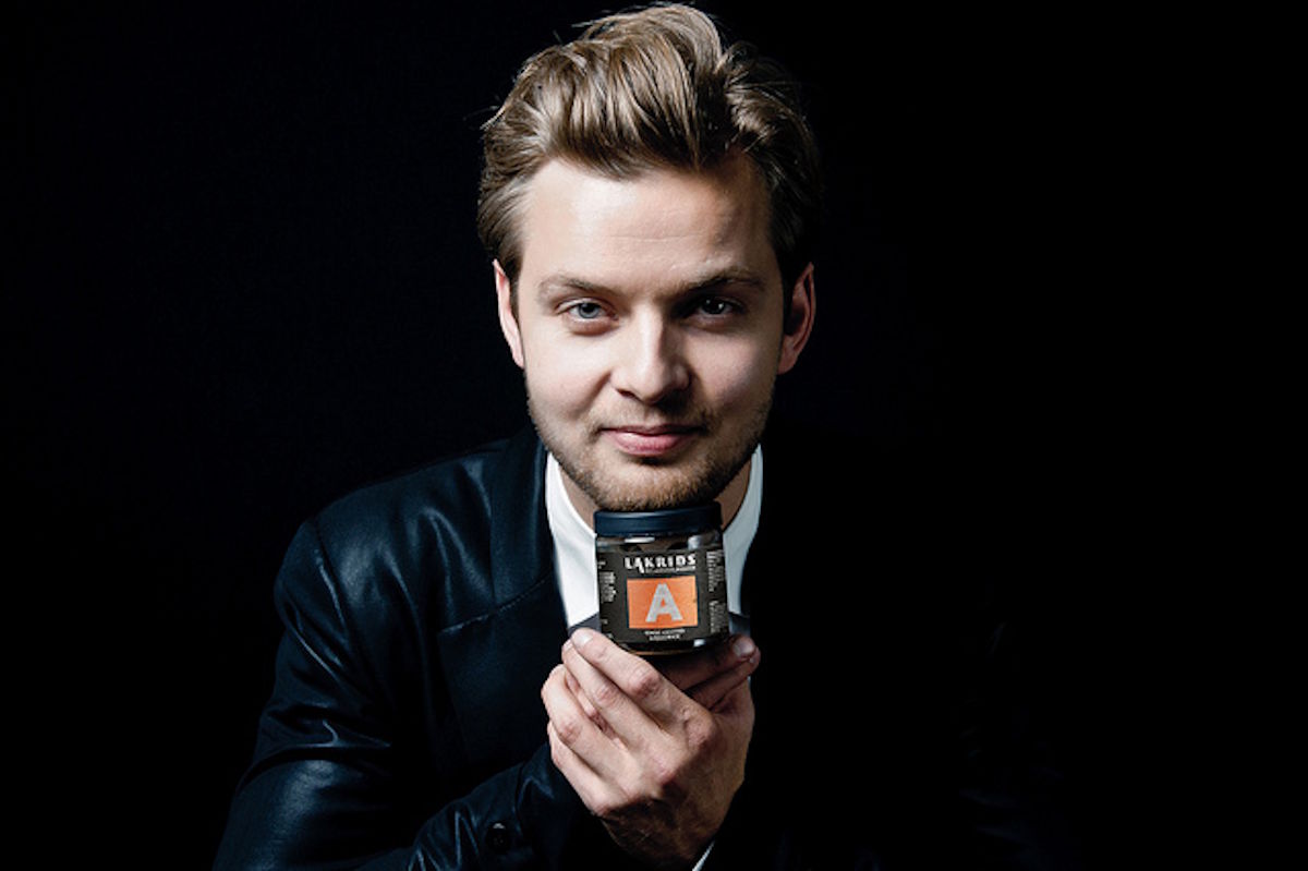 Porträt eines jungen Mannes im dunklen Sakko vor schwarzem Hintergrund, der ein Produktglas mit auffälligem Etikett in den Händen hält und direkt in die Kamera blickt