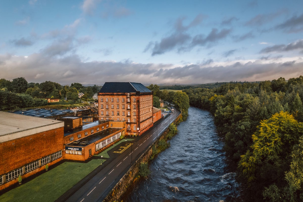 Backsteinfarbenes Industrie- oder Fabrikgebäude mit dunklem Dach direkt an einem schnell fließenden Fluss, umgeben von Wald und Natur unter dramatischem Himmel in den frühen Morgenstunden – möglicherweise Standort einer Brennerei oder Manufaktur