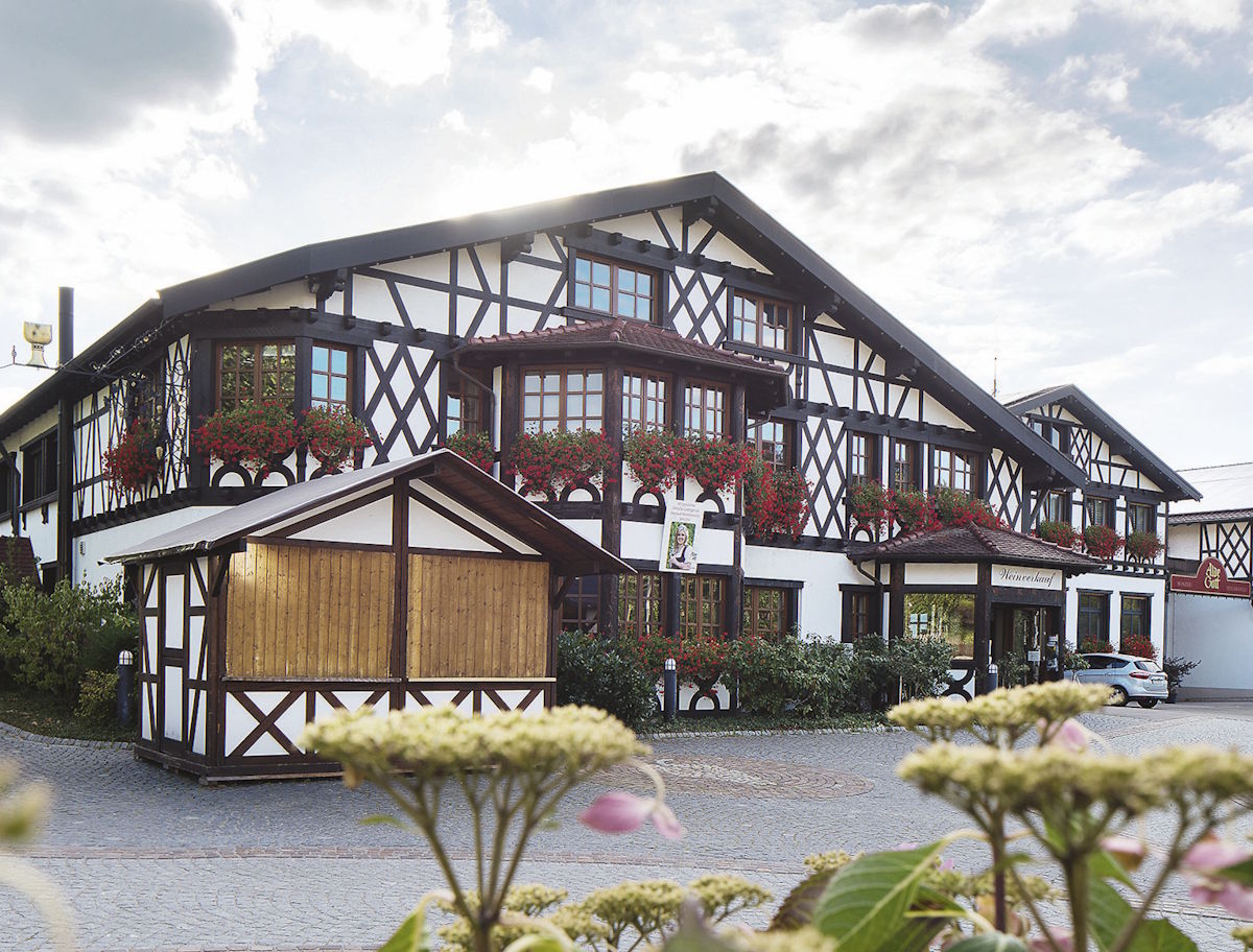Historisches Fachwerk-Weingut mit blumengeschmückten Balkonen, gepflegtem Vorgarten und gepflastertem Innenhof bei sonnigem Wetter