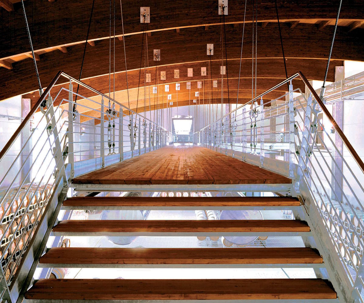 Moderne Brücke mit Holzstufen und Metallgeländer in einem architektonisch gestalteten Weinkeller mit Sicht auf Barriquefässer