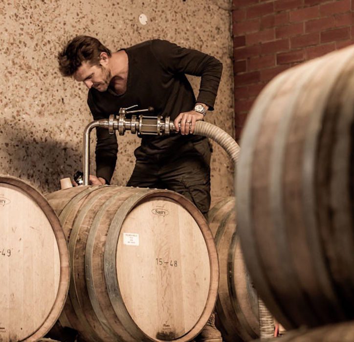 Winzer bei der Arbeit im Weinkeller, füllt Wein mithilfe eines Schlauchs in Holzfässer – umgeben von mehreren Eichenfässern.