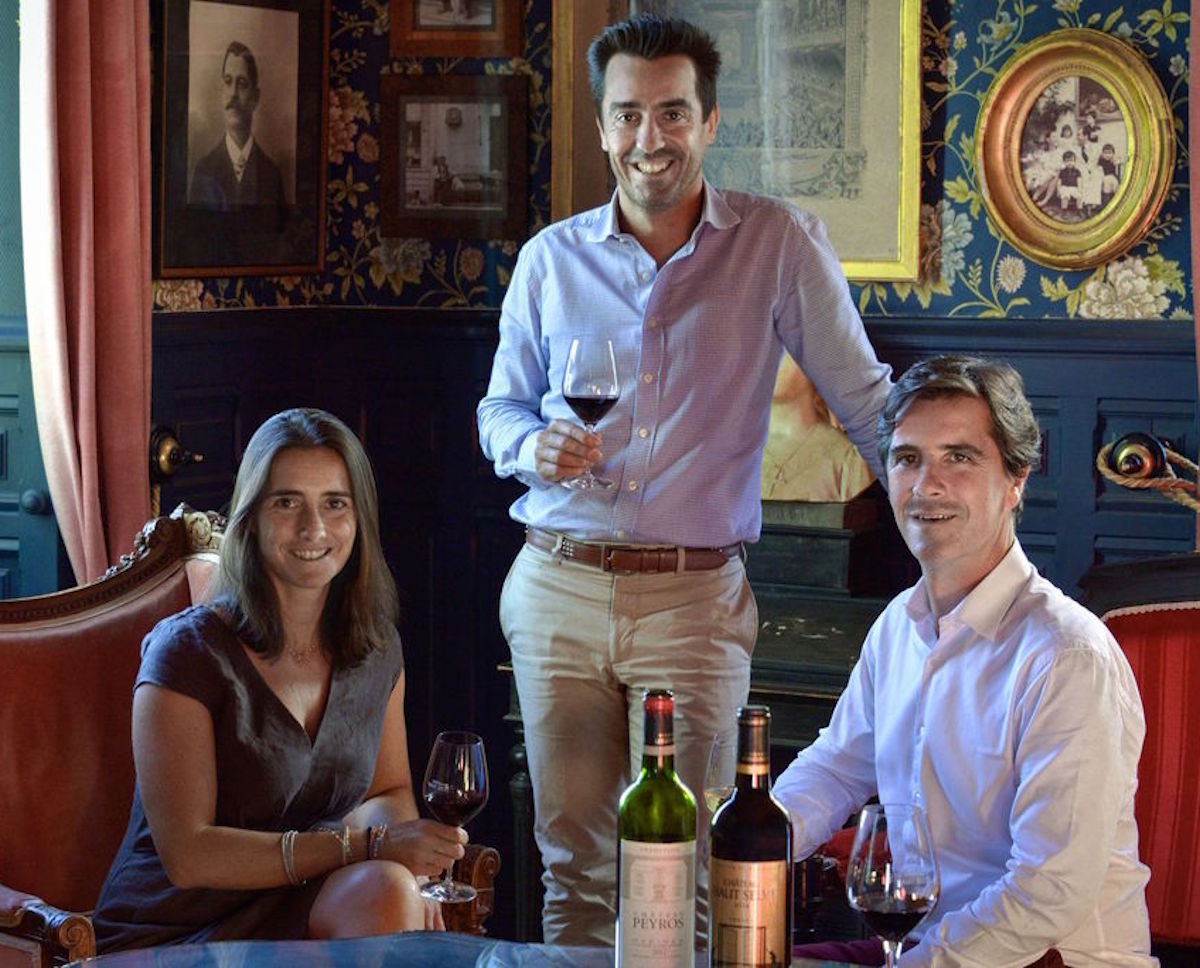 Drei Personen mit Weingläsern in einem elegant eingerichteten Raum mit historischen Porträts und Tapeten, im Vordergrund stehen zwei Weinflaschen auf einem Glastisch.