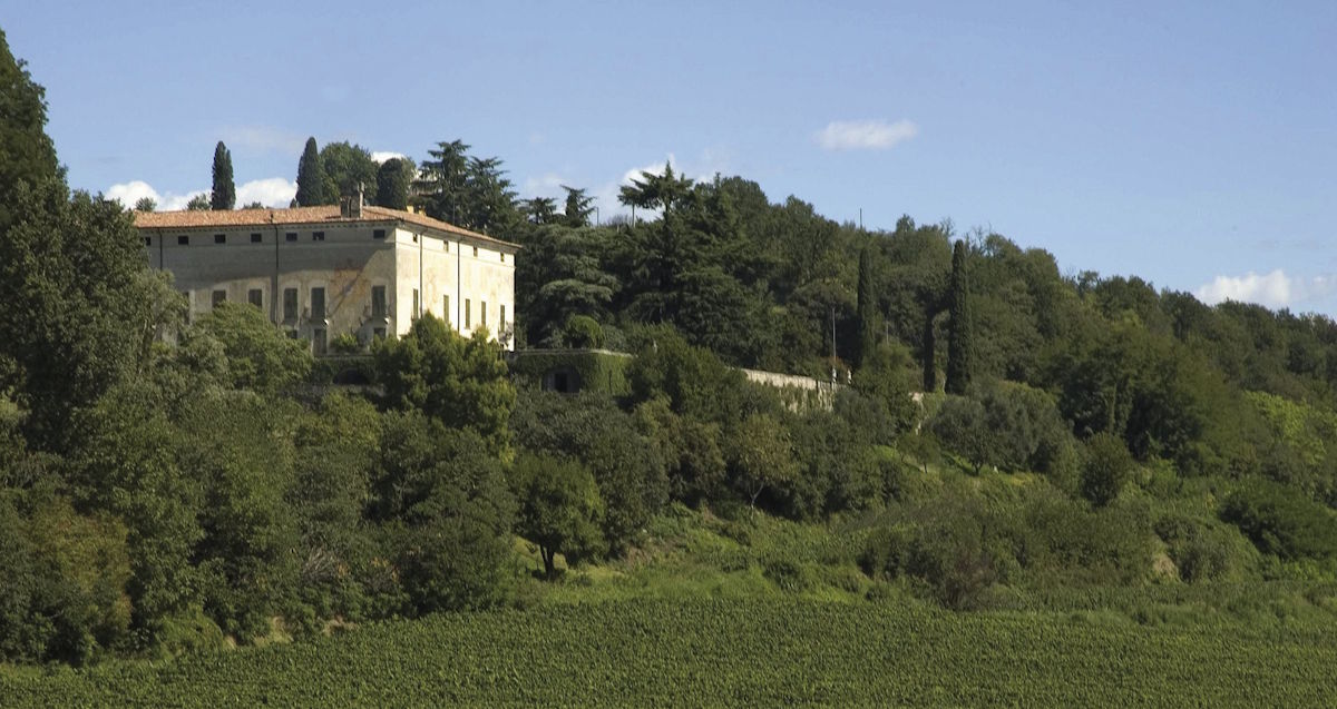 Historisches Gutshaus mit rotem Ziegeldach auf einem Hügel, umgeben von Weinbergen und Zypressen.