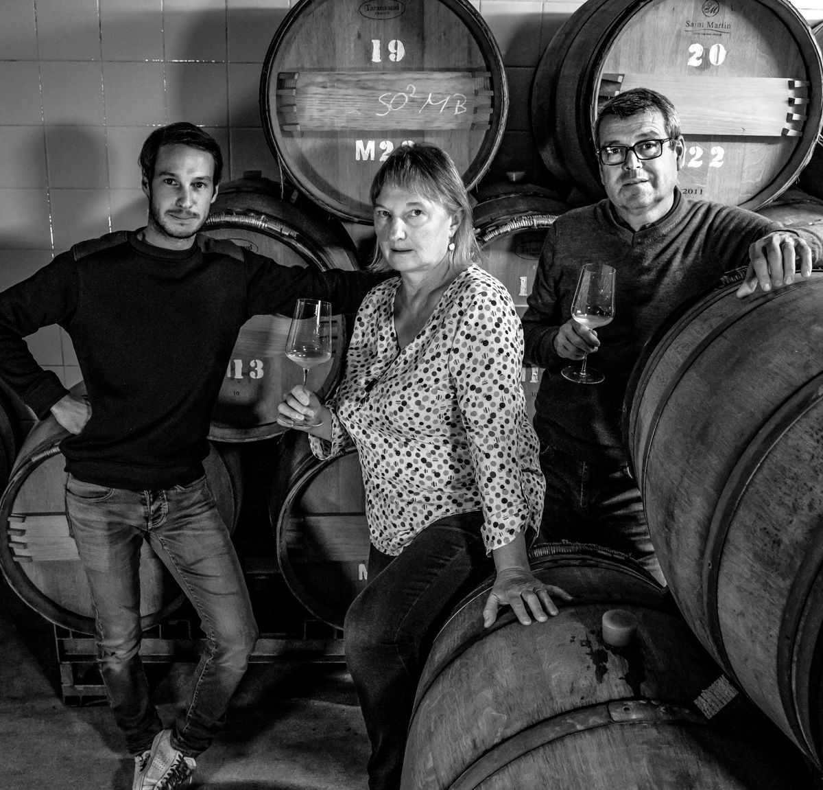 Drei Personen mit Weingläsern posieren zwischen großen Holzfässern in einem Weinkeller, Schwarz-Weiß-Aufnahme.