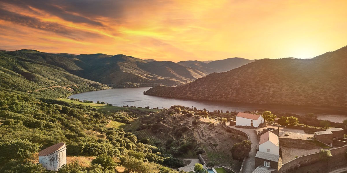 Hügelige Flusslandschaft bei Sonnenuntergang mit kleinen Gebäuden im Vordergrund und bewaldeten Berghängen im Hintergrund.